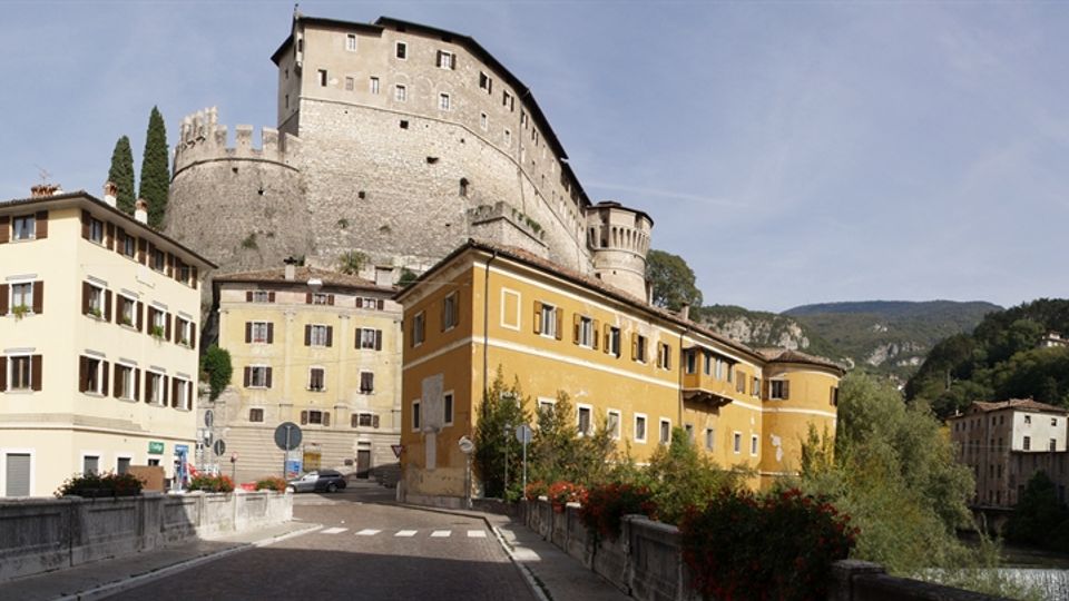 Vista di un borgo con case a più piani affacciate su una strada e un ponte, dominato da un grande castello in pietra con mura e torre circolare sulla collina. Sullo sfondo si vedono colline verdi e un cielo limpido.