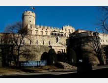 Veduta esterna del Castello del Buonconsiglio, grande complesso fortificato in pietra con torre circolare merlata, mura alte e un ingresso con scalinata e portale classicheggiante. In primo piano si vede un cartello informativo e vegetazione con alberi spogli, sullo sfondo cielo azzurro.