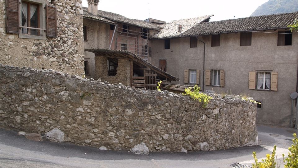 Cortile di un borgo con case in pietra e intonaco, finestre con persiane e tetti in coppi. In primo piano un basso muro curvo in pietra delimita una strada asfaltata, con colline sullo sfondo.
