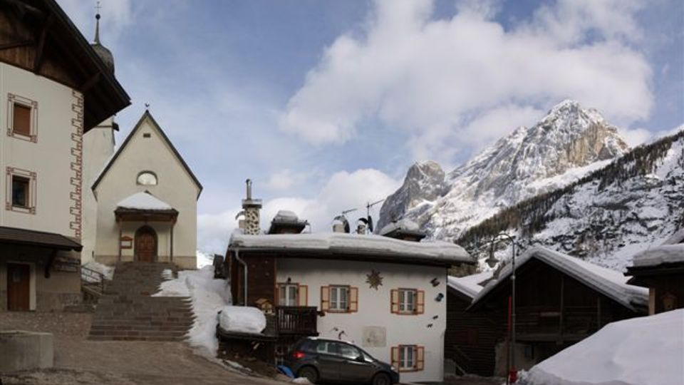 Piccola piazza di un villaggio di montagna innevato con una chiesetta a facciata semplice e scala in pietra, affiancata da case tradizionali con tetti carichi di neve. Sullo sfondo si vedono alte montagne rocciose coperte di neve sotto un cielo parzialmente nuvoloso.