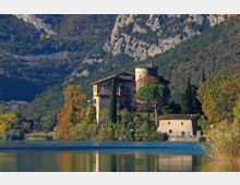 Complesso di edifici storici in pietra con torre cilindrica e tetti in tegole, circondato da cipressi e alberi, sulla riva di un lago. Sullo sfondo si vedono pendii boscosi e alte pareti rocciose di montagna.