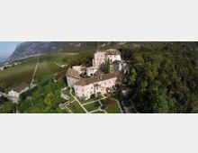 Veduta aerea di un complesso castellano in pietra con tetti in coppi rossi e torre centrale, circondato da giardini geometrici e alberi. Sullo sfondo si vedono vigneti nella valle e montagne rocciose ai margini del bosco.