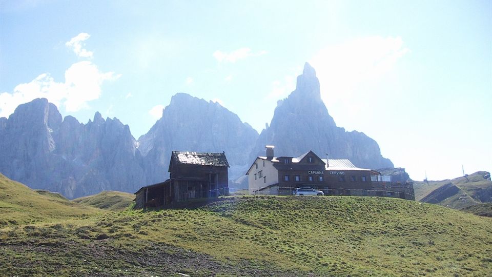Alpeggio di montagna con prati ondulati e alcune baite e un rifugio su un crinale. Sullo sfondo si vedono alte cime rocciose frastagliate contro un cielo luminoso.