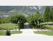 Viale pedonale in un parco curato, con sentiero chiaro fiancheggiato da alberi potati e lampioni in stile classico su prati verdi. Sullo sfondo si vedono campi coltivati e un versante montuoso roccioso.
