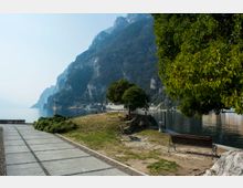 Passeggiata lungolago con pavimentazione in lastre di pietra e una piccola area verde con alberi, panchine e rocce vicino alla riva. Sullo sfondo si vede il lago calmo e un’alta parete montuosa boscosa che domina la costa, con alcuni edifici lontani.
