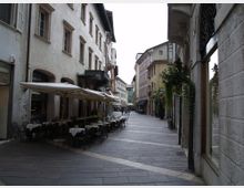 Strada pedonale in un centro storico, pavimentata in pietra e fiancheggiata da edifici alti con facciate intonacate e finestre incorniciate. Sulla sinistra ci sono tavolini di un caffè all’aperto con grandi ombrelloni, mentre a destra si vedono vetrine e piante rampicanti lungo la facciata.
