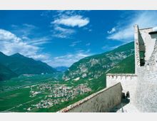 Veduta panoramica da una fortificazione in pietra con mura bianche e camminamento in primo piano, affacciata su una valle con un paese e campi coltivati. Sullo sfondo si vedono alte montagne verdi e rocciose sotto un cielo azzurro con nuvole sparse.