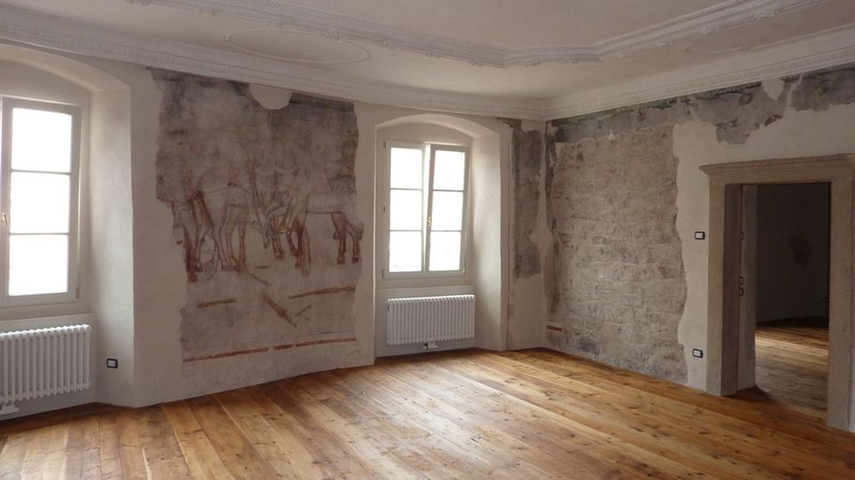 Stanza interna di un edificio storico con pavimento in legno, soffitto decorato con cornici in gesso e due finestre ad arco con radiatori sotto. Sulle pareti sono visibili affreschi parzialmente conservati e porzioni di intonaco rimosso, con un’apertura su un altro ambiente a destra.