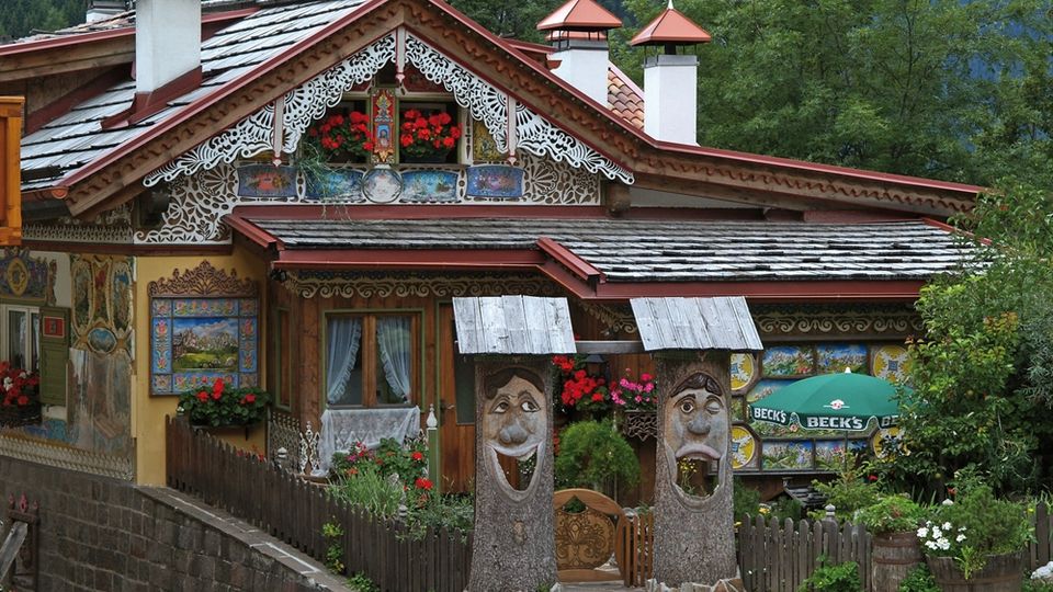 Casa alpina decorata con intagli in legno, motivi dipinti e balconcino con fiori, con tetto a scandole e camini con cappelli rossi. Davanti ci sono una staccionata, un ingresso con due pilastri scolpiti a forma di volti e un piccolo giardino, sullo sfondo boschi e montagne.