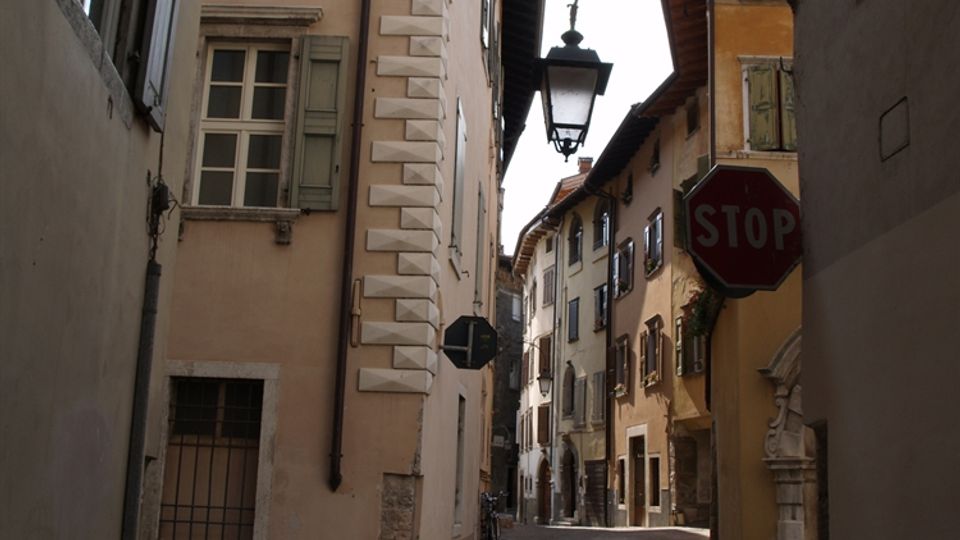 Vicolo stretto con pavimentazione in ciottoli tra alti edifici storici dai colori chiari, con finestre e persiane in legno. Una lanterna sospesa su staffa in ferro battuto e un cartello “STOP” si affacciano sulla strada che curva in lontananza.