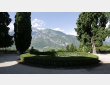 Giardino panoramico con siepi basse potate in forma circolare e vialetti ghiaiosi, incorniciato da alti cipressi. Sullo sfondo si vedono una valle con campi e un’ampia catena montuosa sotto un cielo sereno.