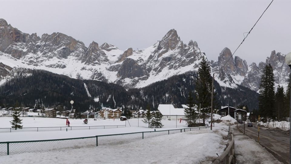 Panorama invernale di una valle con ampi campi innevati e un piccolo centro abitato, sullo sfondo di alte montagne rocciose con cime coperte di neve. In primo piano si vedono una strada con neve ai margini, una recinzione, lampioni e alberi di conifere.