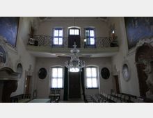 Sala interna storica con soffitti alti, pareti decorate con affreschi e medaglioni, e un grande lampadario di cristallo al centro. In fondo si vedono finestre alte, una porta centrale e una balconata superiore con ringhiera in ferro battuto sopraelevata.
