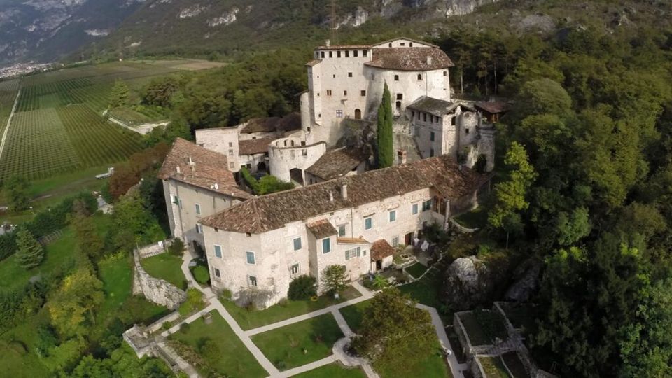 Veduta aerea di un complesso castellano in pietra con tetti in coppi rossi e torre centrale, circondato da giardini geometrici e alberi. Sullo sfondo si vedono vigneti nella valle e montagne rocciose ai margini del bosco.