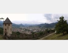 Panorama da una fortificazione in pietra con torre medievale e mura merlate in primo piano, affacciata su una città nella valle. Sullo sfondo si vedono colline e montagne sotto un cielo nuvoloso, con un pendio erboso e alberi lungo il camminamento delle mura.