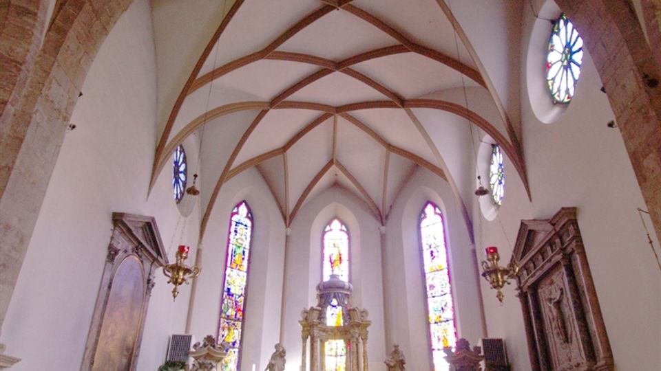 Interno di una chiesa con navata unica e soffitto a volte a costoloni in pietra, che conduce all’altare maggiore rialzato. Sul fondo si vedono alte finestre con vetrate colorate e arredi sacri in stile barocco lungo le pareti laterali.