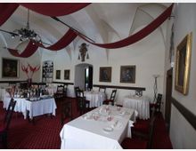 Sala ristorante in stile classico con soffitto a volte bianche e drappi rossi sospesi, lampadario in ferro battuto e pareti decorate con quadri incorniciati. Tavoli apparecchiati con tovaglie bianche su moquette rossa e un’area bar con bottiglie e scaffali sullo sfondo.