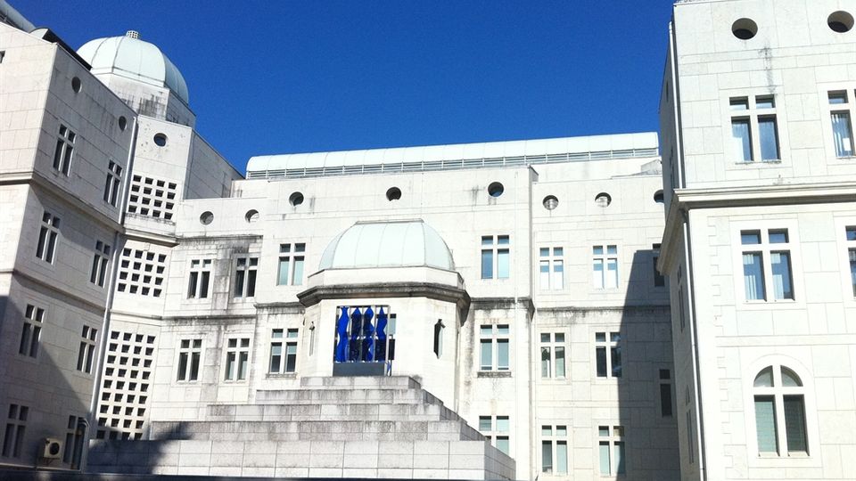 Facciata di un grande edificio istituzionale in pietra chiara, con molte finestre rettangolari e aperture circolari, e piccole cupole sul tetto. Al centro sporge un volume a gradoni con una cupola e una vetrata blu, sotto un cielo limpido.