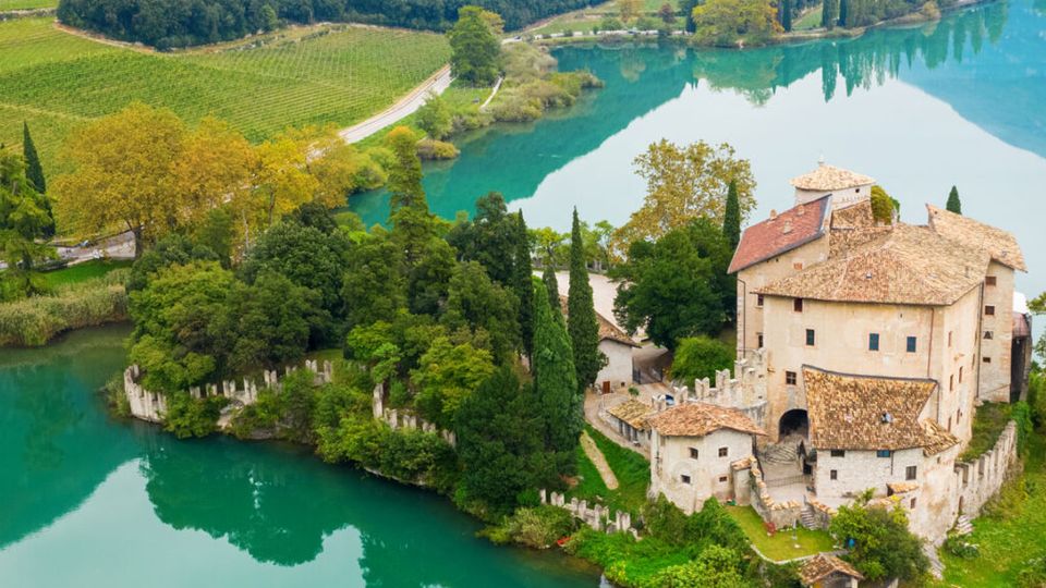 Veduta aerea di un castello in pietra con tetti in coppi, circondato da mura e cipressi su una piccola penisola. L’edificio si affaccia su un lago turchese, con vigneti e strade di campagna sullo sfondo.