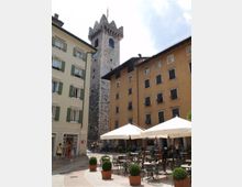 Piazza urbana con caffè all’aperto, tavolini e ombrelloni bianchi, circondata da edifici storici con facciate color ocra e chiara e finestre con persiane. Sullo sfondo si alza una torre medievale in pietra con merlature e piccole finestre sotto un cielo parzialmente nuvoloso.