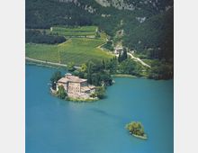 Vista dall’alto di un castello in pietra su una piccola penisola che si protende in un lago turchese, circondato da alberi e cipressi. Sullo sfondo si vedono vigneti terrazzati e un pendio boscoso ai piedi di una parete rocciosa.