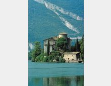 Edificio storico in pietra con torre rotonda e tetti in coppi, circondato da cipressi e alberi sulla riva di un lago. Sullo sfondo si vedono alte montagne boscose con una fascia di roccia chiara.