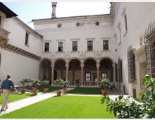 Cortile interno di un palazzo storico con prato verde attraversato da vialetti e vasi di agrumi. Sul fondo si vede un loggiato ad archi sostenuti da colonne e una facciata chiara con finestre e piccoli oculi.