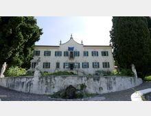 Facciata di una villa storica intonacata di bianco con persiane verdi e frontone centrale, affacciata su un giardino rialzato con muretto e piccola fontana. Due grandi alberi ai lati incorniciano l’edificio, con statue in pietra lungo il bordo del giardino.