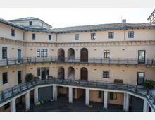 Cortile interno di un edificio storico a più piani con facciate intonacate, arcate e colonne al piano terra. Ballatoi con ringhiere metalliche collegano finestre e porte, con una scala centrale che sale ai livelli superiori sotto un tetto in tegole.