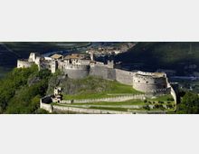 Veduta aerea di un grande castello medievale in pietra con bastioni, torri e mura merlate, disposto su più livelli lungo una collina boscosa. In primo piano si vedono le fortificazioni esterne e un prato interno, con la valle e un centro abitato sullo sfondo.