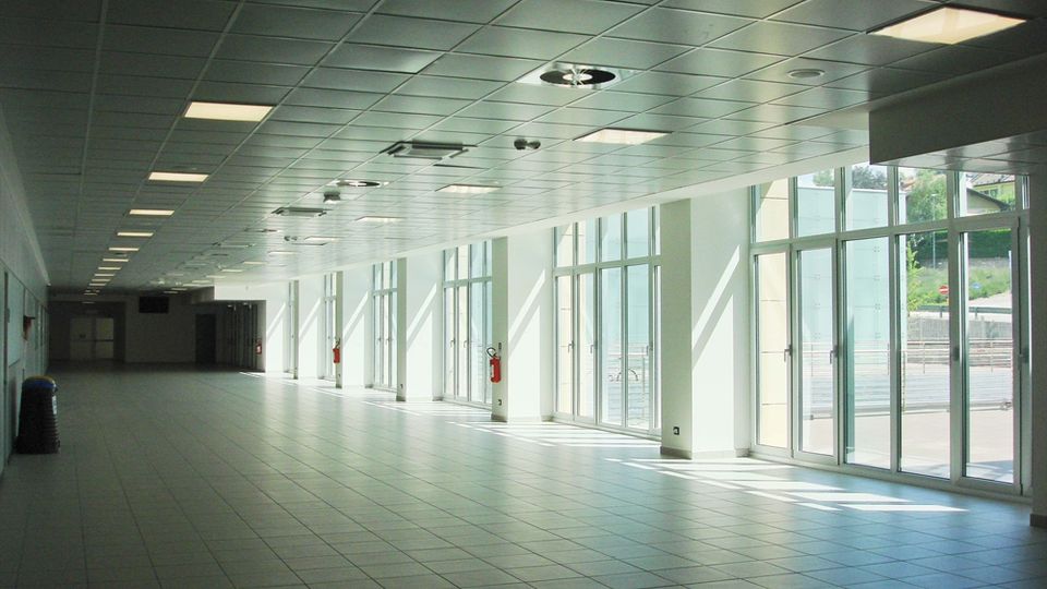 Ampio corridoio interno moderno con pavimento piastrellato e controsoffitto a griglia con pannelli luminosi. Lungo un lato ci sono grandi vetrate e porte a tutta altezza che lasciano entrare luce naturale e affacciano su un’area esterna.