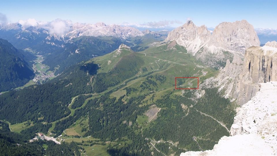 Veduta panoramica di una valle alpina con boschi e prati attraversati da strade e sentieri, con un piccolo centro abitato sul fondo. Sullo sfondo si elevano imponenti cime rocciose e pareti montuose sotto un cielo parzialmente nuvoloso.