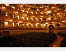 Interno di un teatro all’italiana con platea di poltrone scure e più ordini di palchi curvi sovrapposti. Pareti e balconate decorate in stile classico, illuminate da numerose lampade calde, con un ingresso ad arco sul fondo.