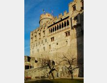 Facciata di un castello in pietra con mura merlate e una torre cilindrica sormontata da bandiere. In alto si vede una loggia con archi e colonne, mentre in basso ci sono un cortile con alberi spogli, panchine e un muretto in pietra sotto un cielo sereno.