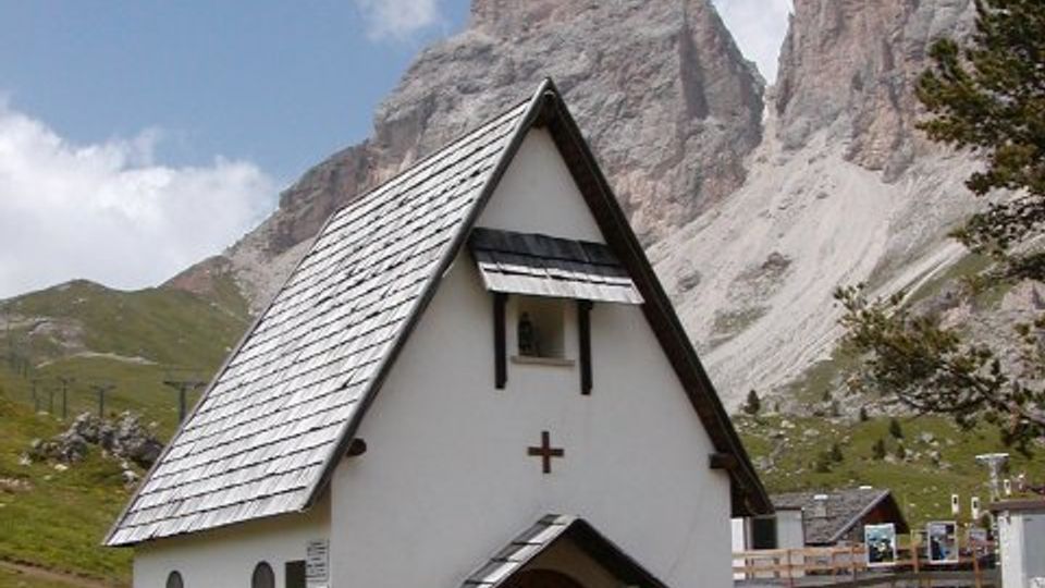 Piccola cappella di montagna bianca con tetto molto spiovente in scandole e portale in legno, su un prato alpino. Sullo sfondo si vedono alte cime rocciose e pendii verdi sotto un cielo parzialmente nuvoloso.