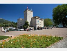 Castello in pietra con alta torre merlata affacciato su un porto con barche ormeggiate e un lungolago. In primo piano una piazza lastricata con aiuola fiorita, alberi e montagne sullo sfondo sotto cielo sereno.