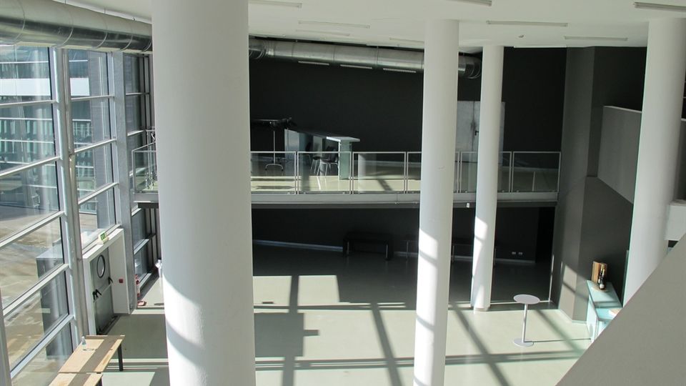 Atrio interno moderno a doppia altezza con grandi vetrate laterali, colonne cilindriche bianche e soppalco con parapetto in vetro. Pavimento chiaro con ombre geometriche della luce e pareti scure con condotte a vista lungo il soffitto.