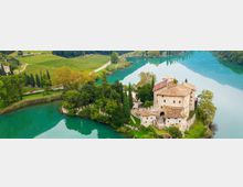 Veduta aerea di un castello in pietra con tetti in coppi, circondato da mura e cipressi su una piccola penisola. L’edificio si affaccia su un lago turchese, con vigneti e strade di campagna sullo sfondo.