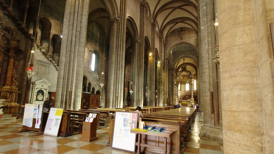 Interno di una grande chiesa in stile gotico, con navata centrale lunga, alte colonne in pietra e volte a crociera. Panche in legno disposte in file e pavimento a scacchi conducono verso l’altare illuminato in fondo.