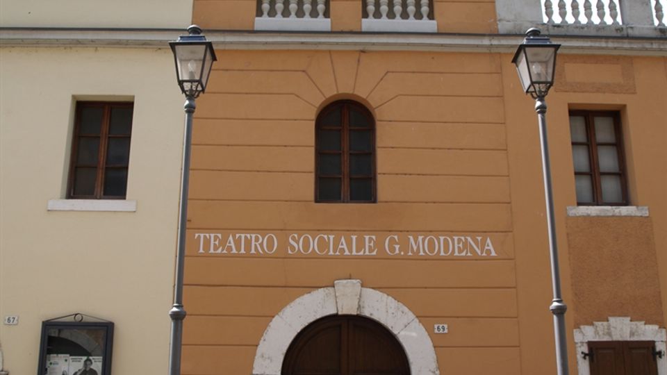 Facciata di un teatro storico con intonaco ocra e bianco, ingresso ad arco in pietra e portone in legno; sopra l’entrata si legge “TEATRO SOCIALE G. MODENA”. Due lampioni in ferro incorniciano l’accesso e una piccola torre centrale mostra finestre ad arco e una balaustra.