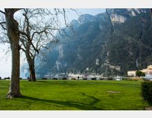 Parco sul lungolago con ampio prato verde, alberi spogli e panchine lungo la riva. Sullo sfondo si vedono un molo con barche e edifici bassi ai piedi di alte montagne rocciose e boscose.