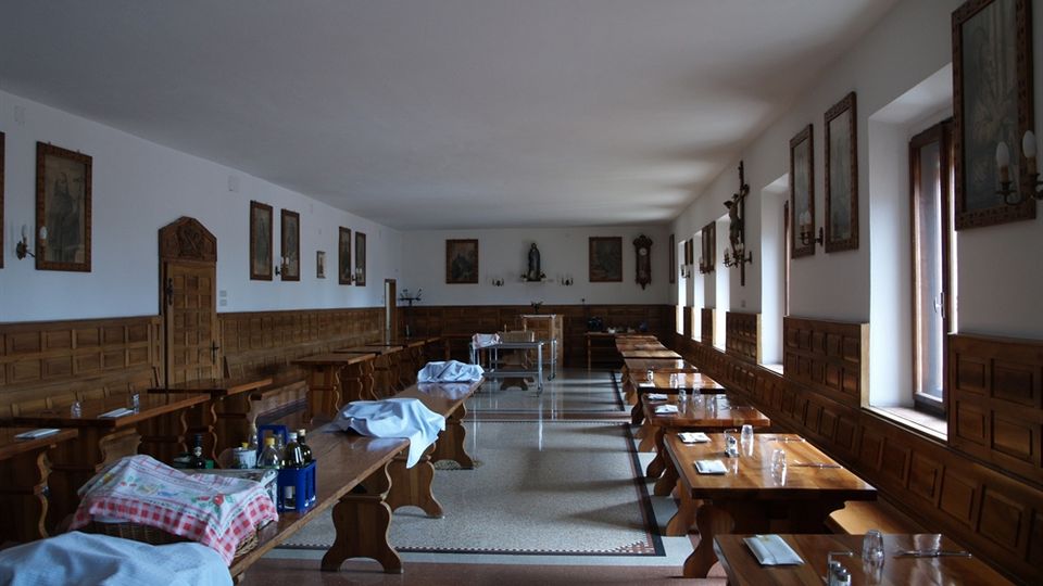 Sala da pranzo lunga con pannellature in legno alle pareti, file di tavoli e panche in legno apparecchiati e pavimento a mosaico. Grandi finestre sul lato destro illuminano l’ambiente, con quadri e arredi tradizionali alle pareti.