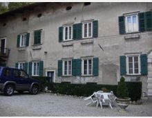 Facciata di un edificio residenziale in intonaco chiaro, a due piani, con numerose finestre dotate di persiane verdi e piccole mensole sotto i davanzali. Davanti c’è un cortile in ghiaia con siepi basse, un grande vaso ornamentale e un gruppo di sedie e tavolino in plastica.