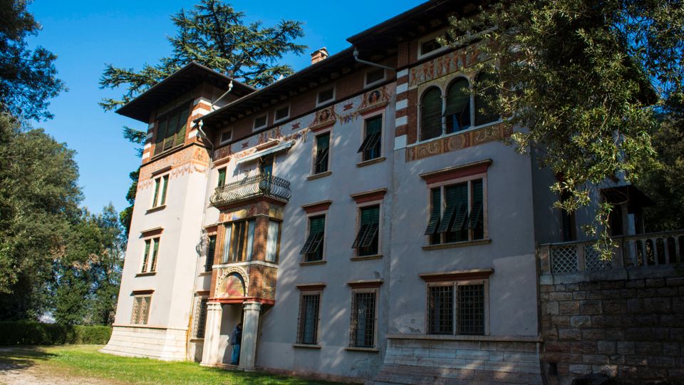 Facciata di una villa storica a più piani con intonaco chiaro, decorazioni pittoriche e cornici in mattoni, finestre con persiane verdi e balconcino centrale sopra un portale ad arco. L’edificio è circondato da prato e alberi alti, con un muretto in pietra sul lato destro.
