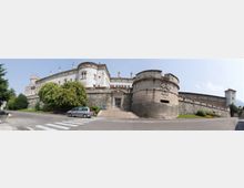 Veduta panoramica di una grande fortezza in pietra su un lieve pendio, con mura alte e un massiccio bastione cilindrico in primo piano. Davanti si vedono una strada con strisce pedonali, un’auto parcheggiata e alberi e aiuole lungo il perimetro delle fortificazioni.