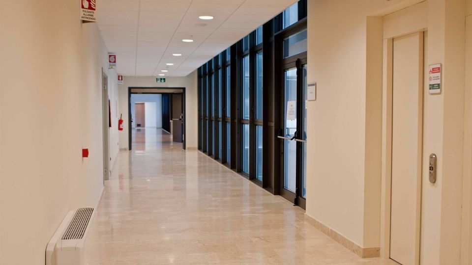 Corridoio interno moderno con pavimento lucido in marmo chiaro, soffitto con pannelli e luci incassate. Sul lato destro una lunga parete di finestre e una porta vetrata, con porte e segnaletica di sicurezza lungo le pareti.