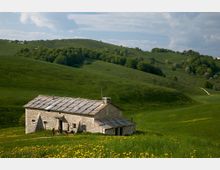 Casa rurale in pietra con tetto a lastre grigie su un prato verde ondulato, circondata da colline e boschi. Sullo sfondo si vedono pendii erbosi, un sentiero lontano e cielo parzialmente nuvoloso.