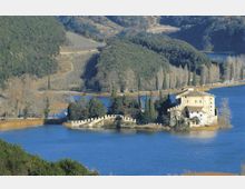 Castello in pietra su un piccolo isolotto in un lago, collegato alla riva da un ponte arcuato con parapetti merlati. Sullo sfondo si vedono colline boscose, filari di cipressi lungo la sponda e campi coltivati.