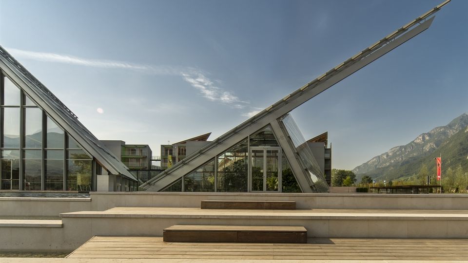 Edificio moderno con grandi vetrate e due elementi architettonici inclinati a forma di lama che si incrociano sopra una terrazza a gradoni con panche in legno. Sullo sfondo si vedono altri edifici contemporanei e una catena montuosa sotto un cielo sereno.