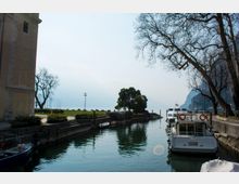 Canale portuale che sfocia in un lago, con barche ormeggiate lungo banchine in pietra e un’imbarcazione più grande sulla destra. Sulla riva si vedono alberi spogli, un edificio sul lato sinistro, un piccolo promontorio alberato e montagne sullo sfondo.